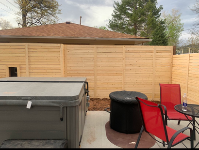 Backyard patio with hot tub, red chairs, round black fire pit, and wooden privacy fence.