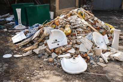 A large pile of construction debris and broken bathroom sinks sitting outdoors next to two green trash bins.