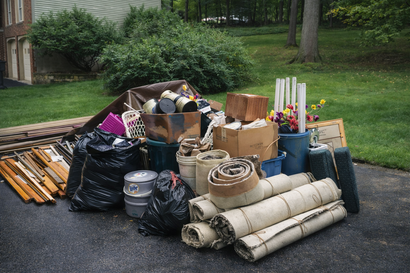 A pile of household junk, including trash bags, rolled rugs, a wooden box, and building materials, sits on a driveway.