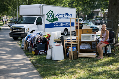 A woman sits in a chair next to a collection of household items and a Lowry Place moving truck parked on a grass verge.