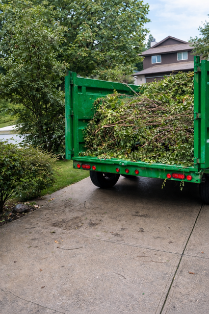 A green dump truck trailer parked on a concrete driveway, filled with yard trimmings and brush in front of a house.