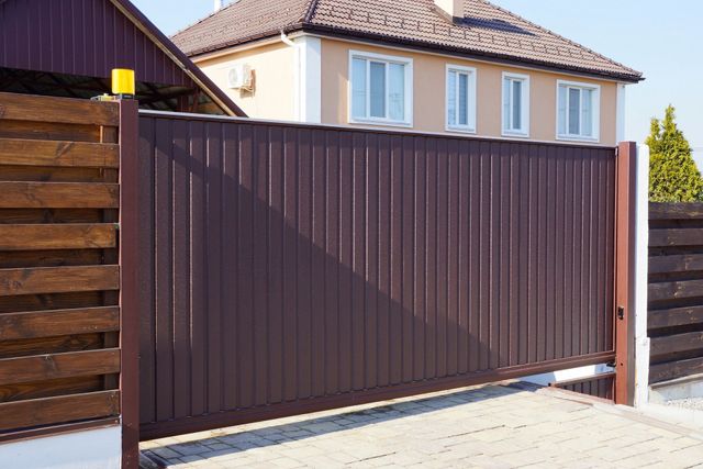 Un cancello scorrevole in metallo marrone installato davanti a una casa residenziale con facciata color beige e tetto marrone scuro.