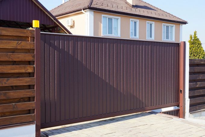 Un cancello scorrevole in lamiera ondulata marrone si erge davanti a una casa, fiancheggiato da sezioni di recinzione in legno.