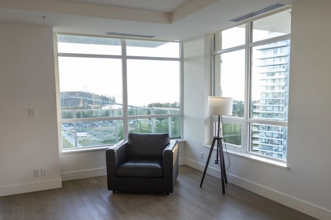 Una poltrona grigio scuro e una lampada da terra a treppiede si trovano in una stanza luminosa e moderna con ampie finestre che si affacciano su una vista della città.