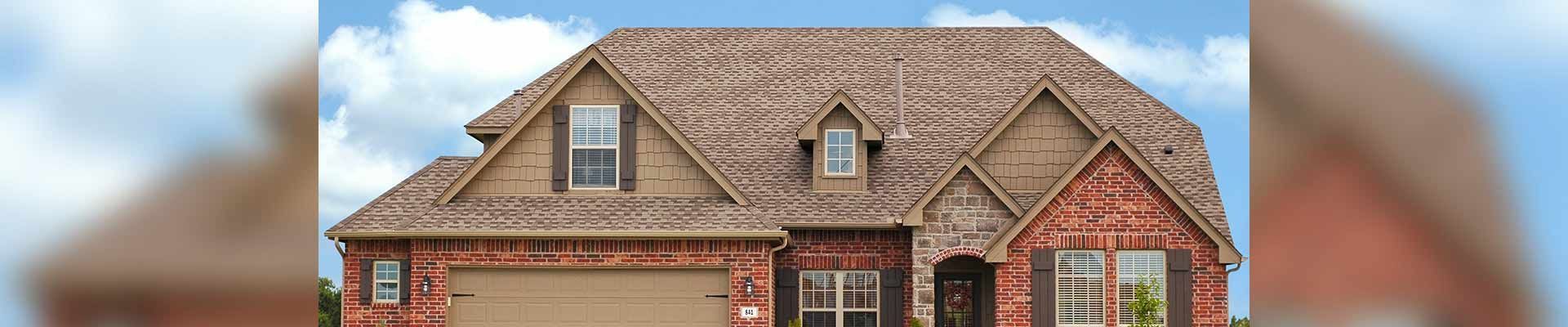 A two-story brick home with a tiled roof. A garage door and several windows are visible. A cloudy blue sky.