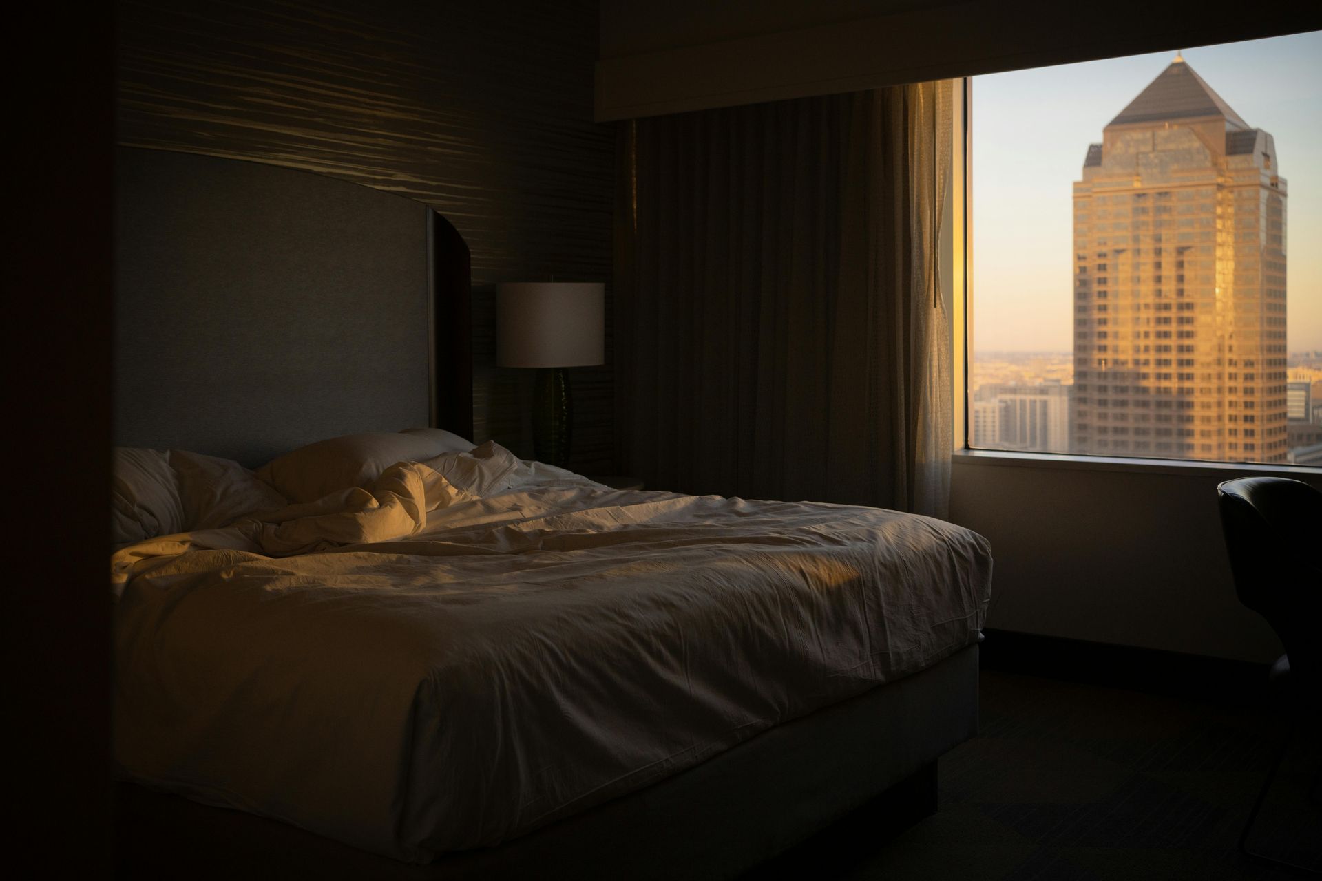 Bedroom with unmade bed; sunlight streams through window, illuminating a skyscraper.