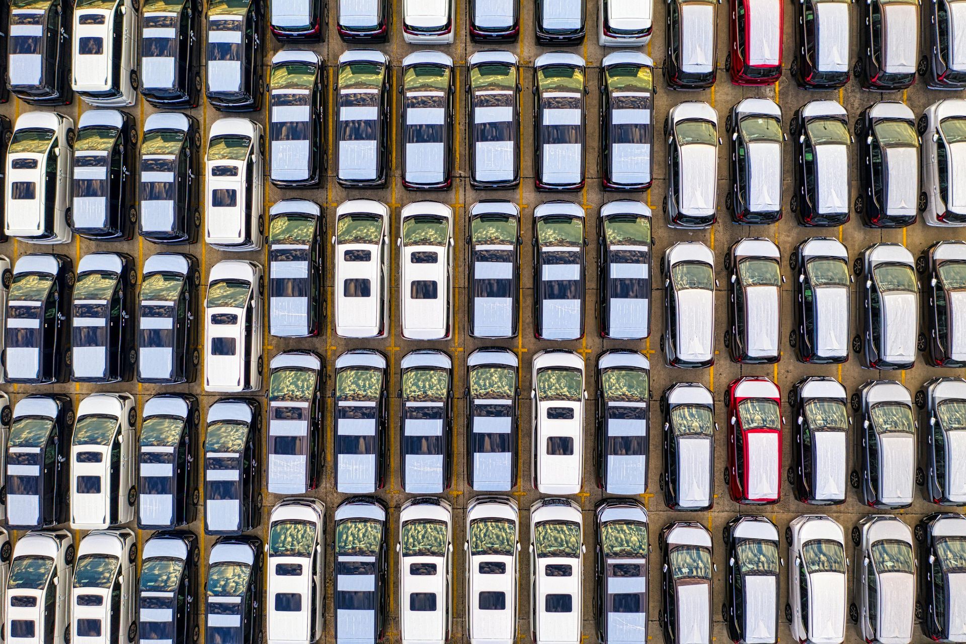 Overhead view of many parked cars in rows. Mostly silver and white vehicles. Two red cars are visible.