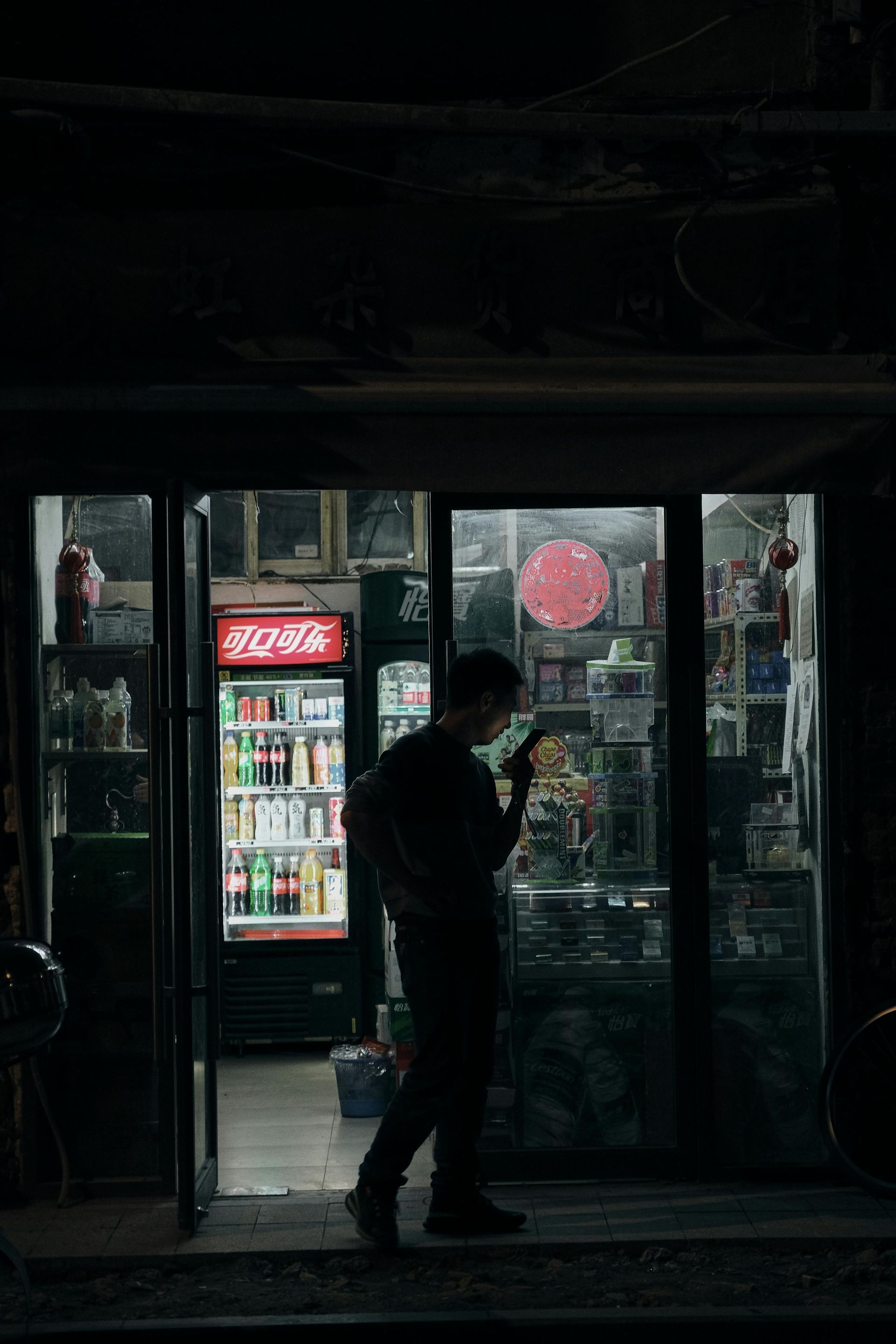 Man outside convenience store at night, smoking. Lit store with red sign, drinks in cooler.