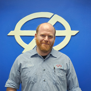 Man with red beard, wearing a light blue work shirt, stands in front of a blue wall with a white logo.