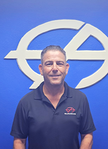 Man in black polo shirt smiles in front of a blue wall with a white logo.
