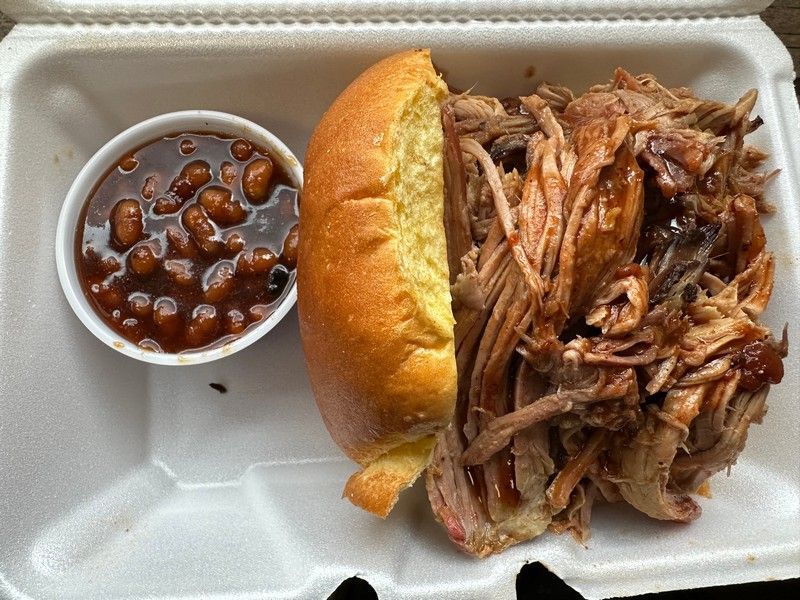 A sandwich with pulled pork and baked beans in a styrofoam container.