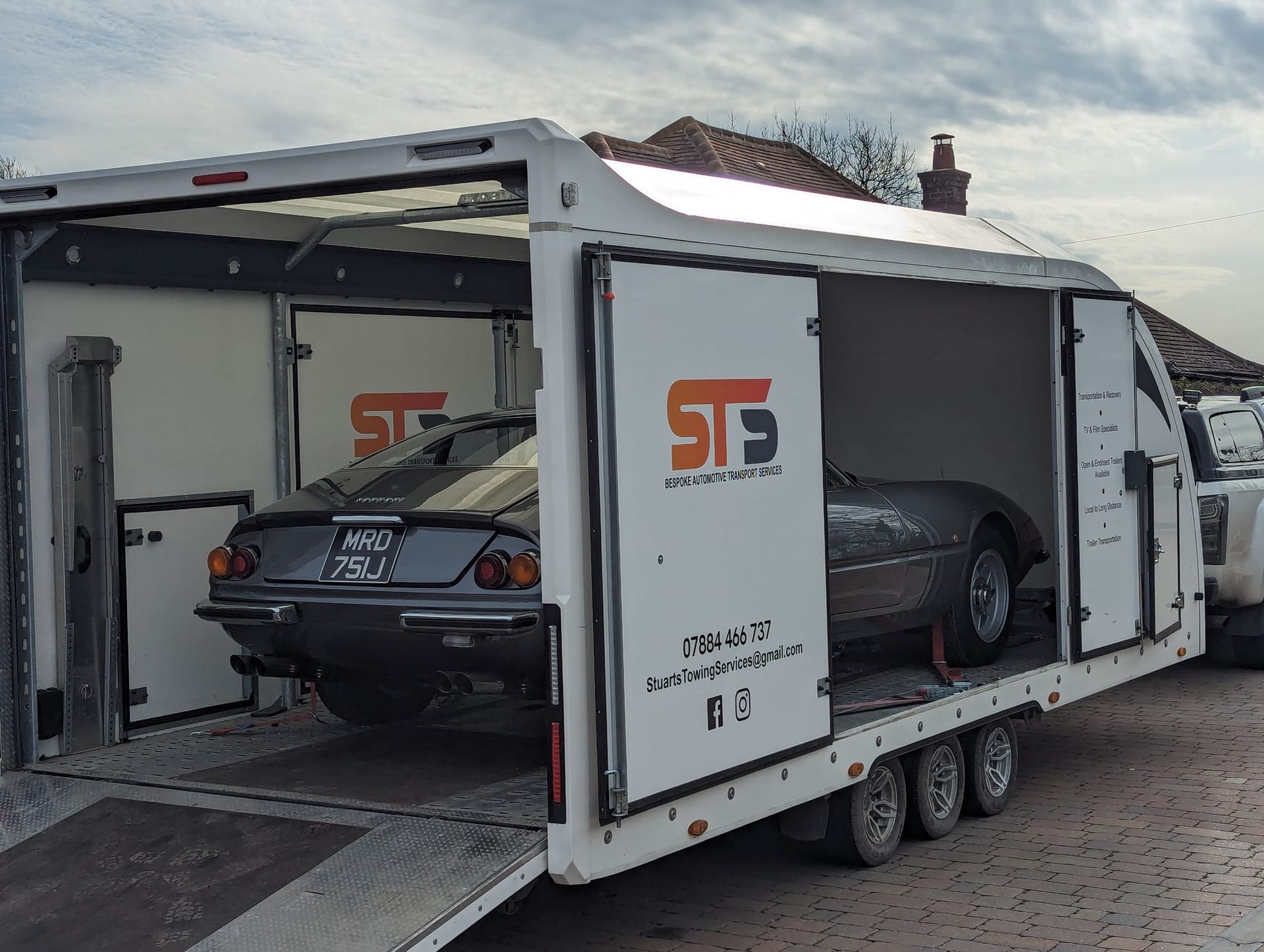 Classic car in enclosed trailer, doors open, being loaded. Gray car, white trailer with logo, outdoor setting.