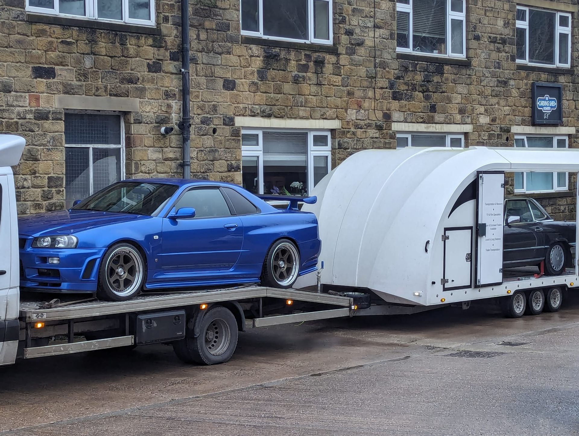 Blue Nissan Skyline R34 on a car transporter, alongside a black car inside. Exterior shot.