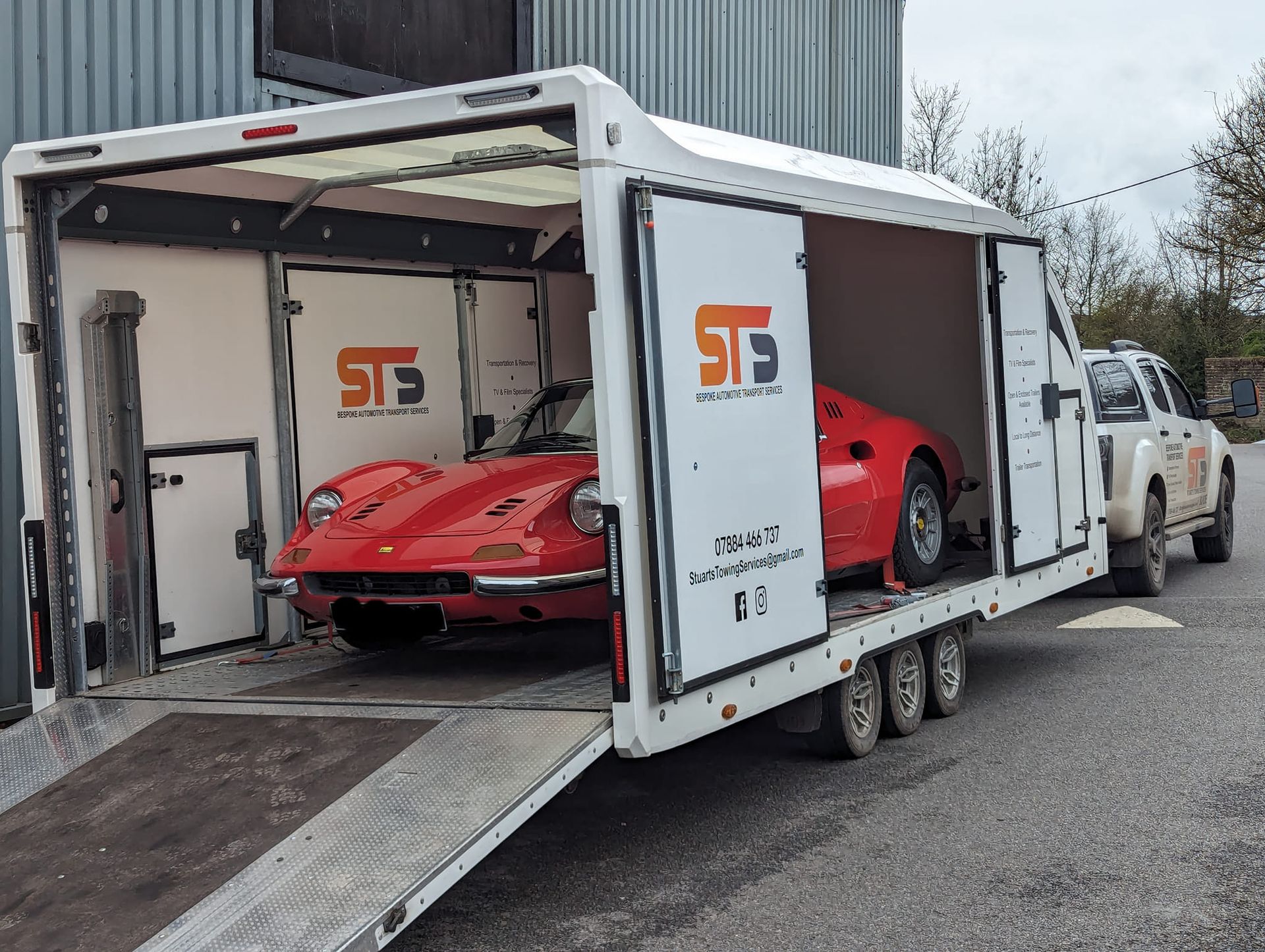 Red classic car being loaded into a white enclosed car trailer by a white truck outdoors.
