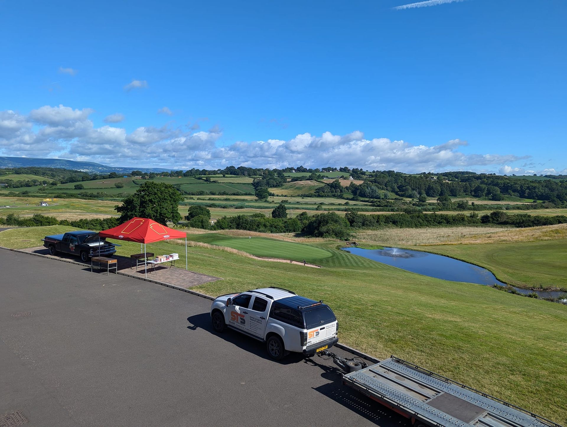 Two white trucks with trailers parked on asphalt overlooking a green landscape with a pond and blue sky.