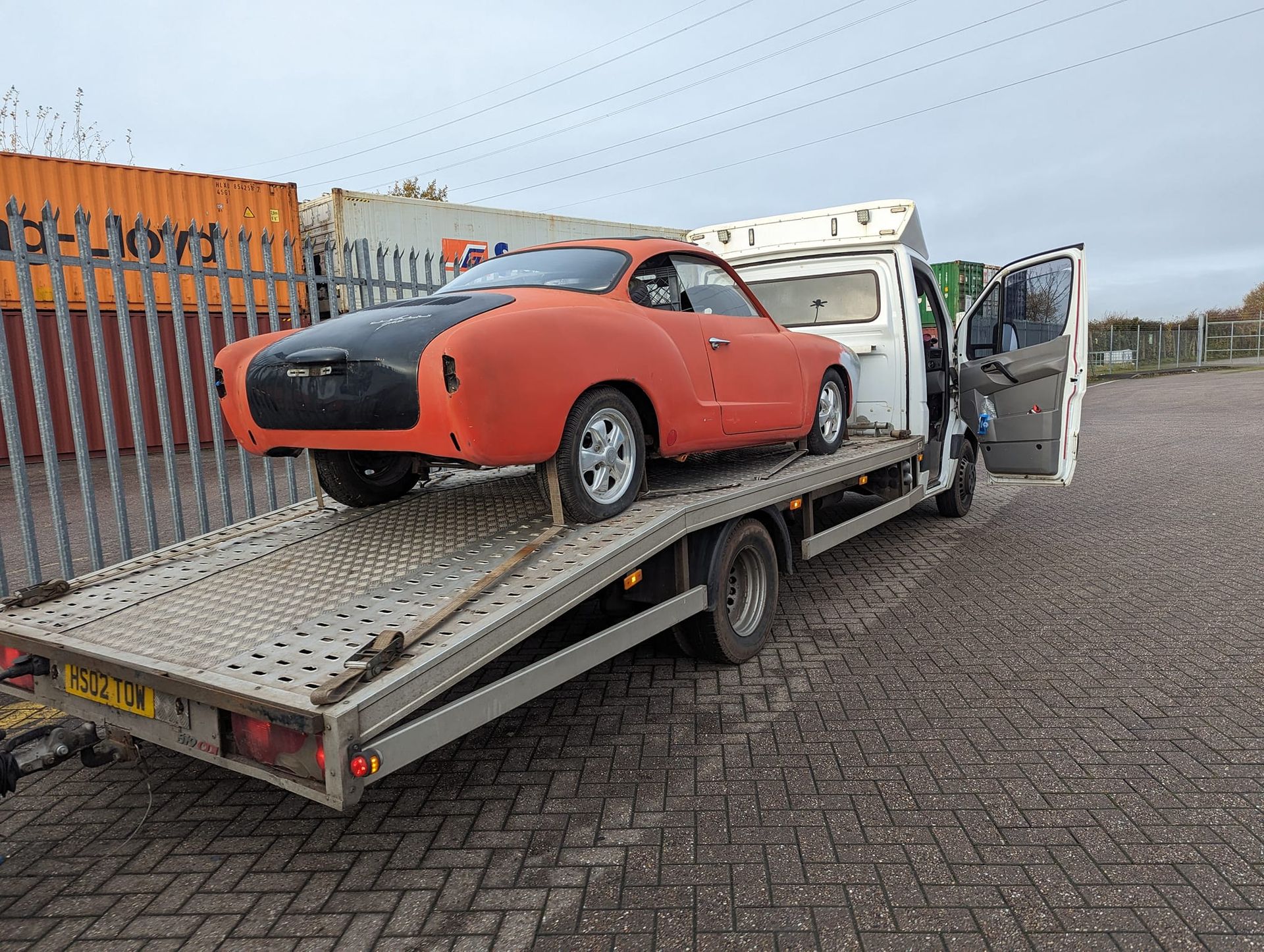 Orange classic car on a flatbed truck, being transported in an industrial area.
