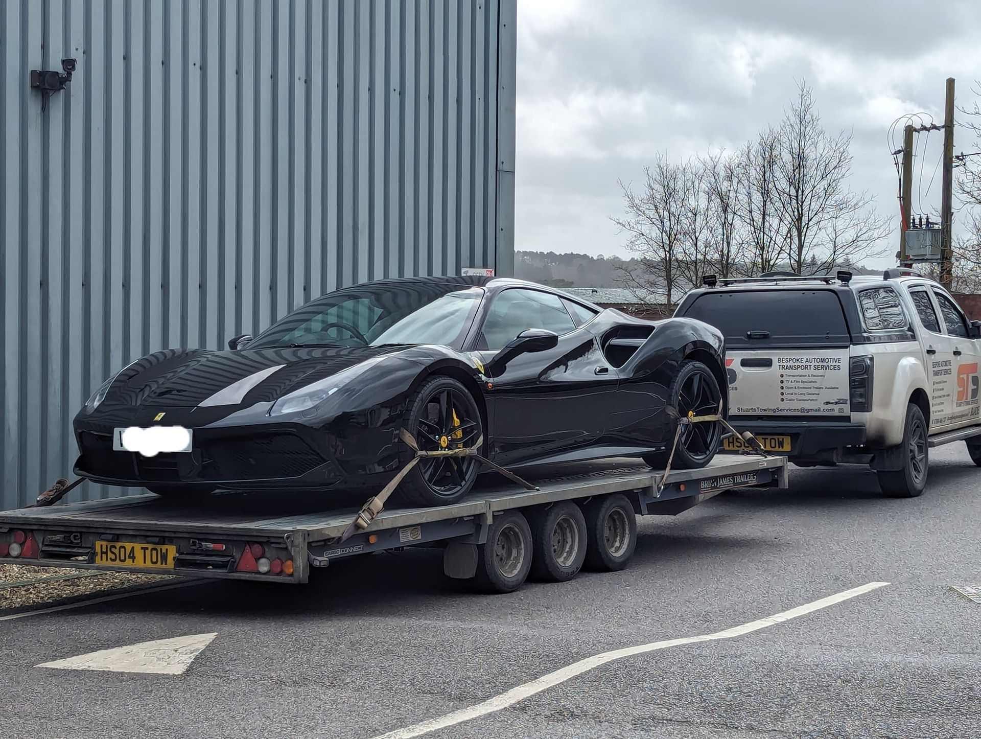 Black Ferrari sports car on a trailer, towed by a white pickup truck on a gray road.