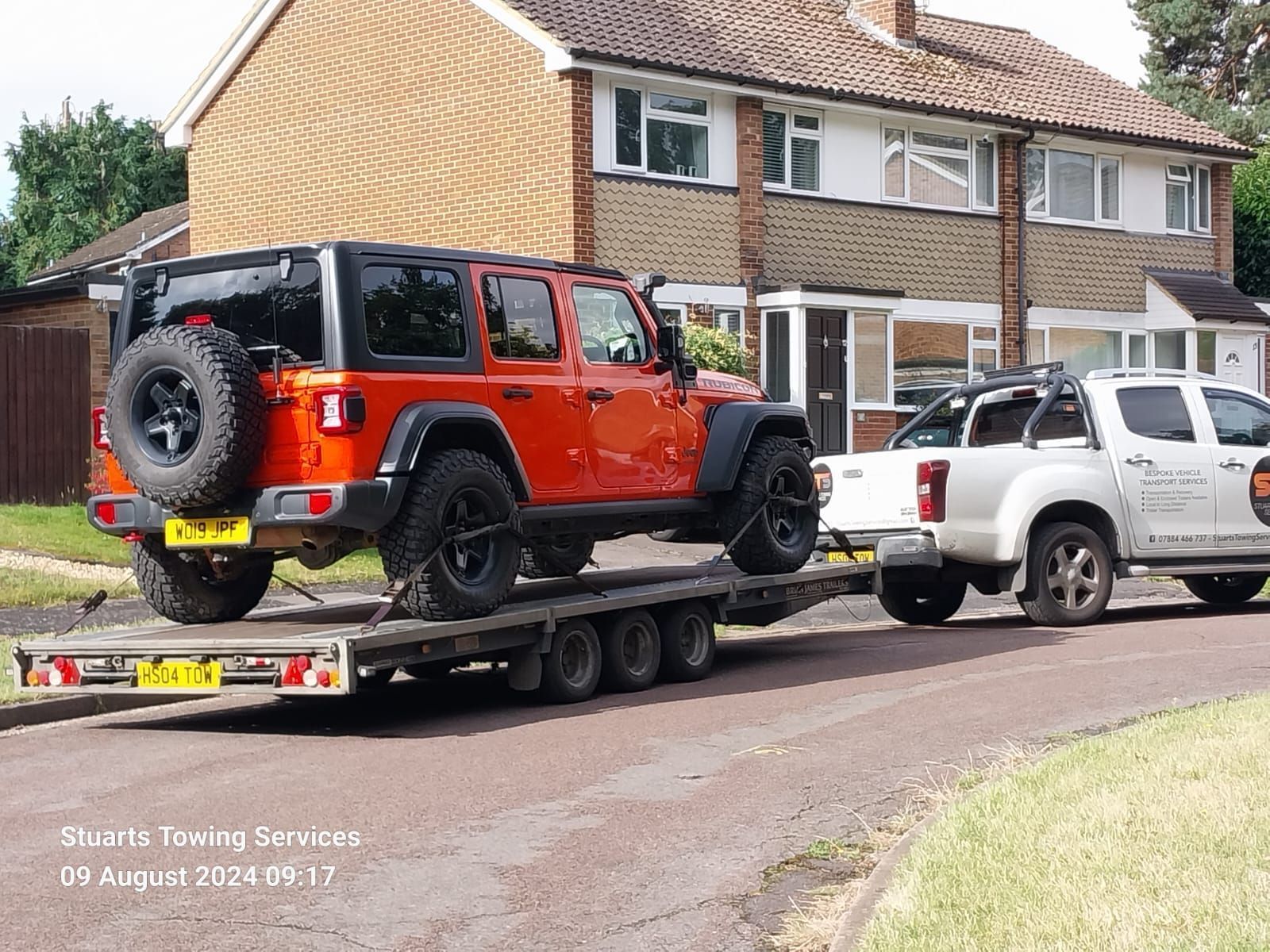 Orange Jeep Wrangler on a trailer being towed by a white pickup truck in front of a residential house.