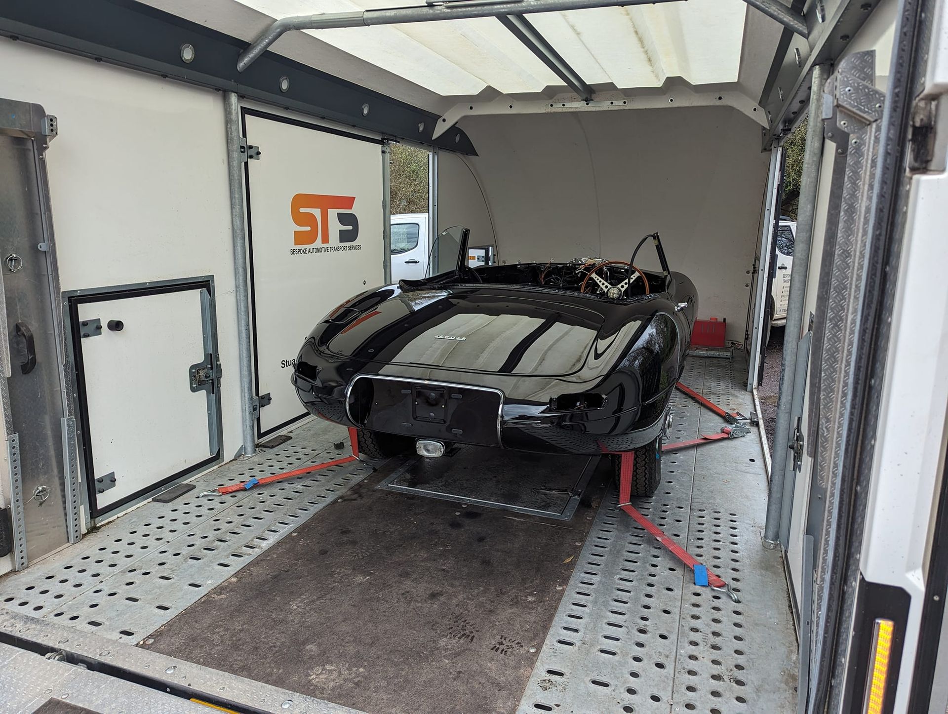 Black vintage car inside a transport trailer, secured with straps.