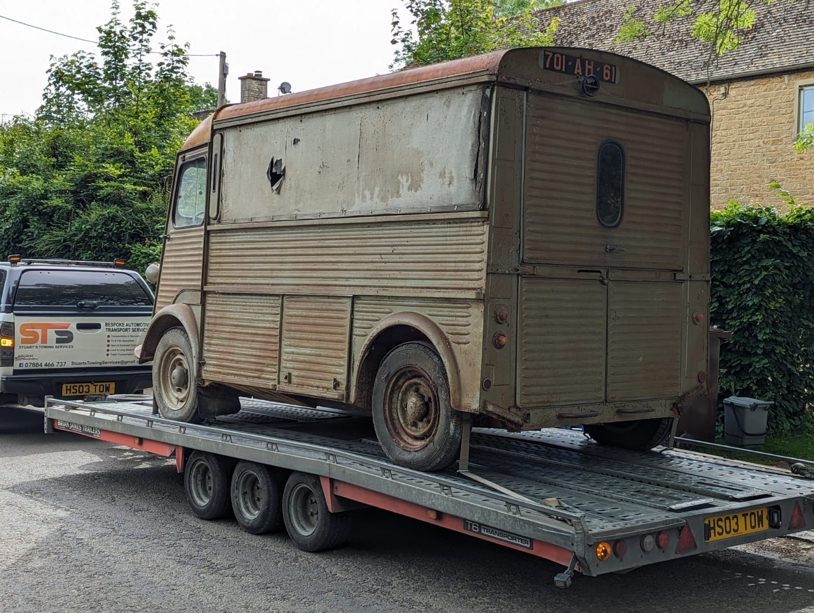 An old, rusty Citroen H van is on a flatbed trailer, being transported on a road.