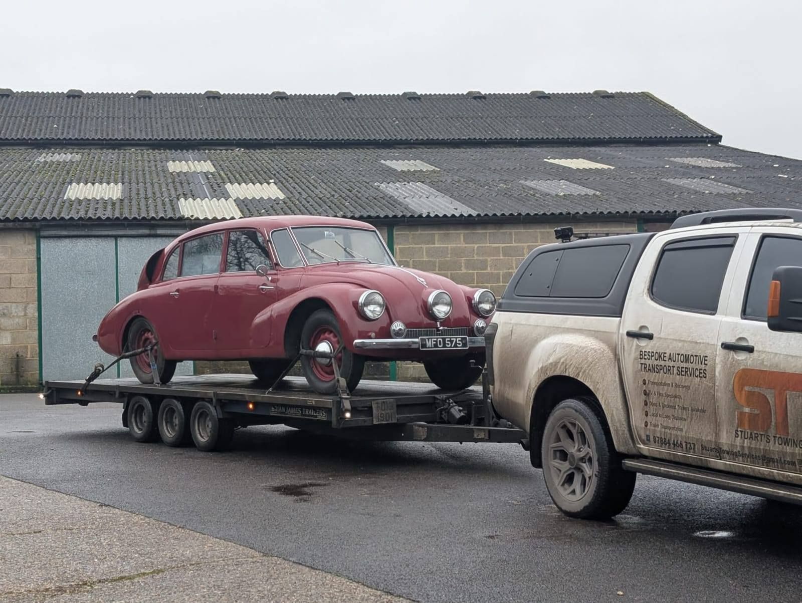 Red classic car on a trailer being towed by a white truck. Outdoors, overcast sky.