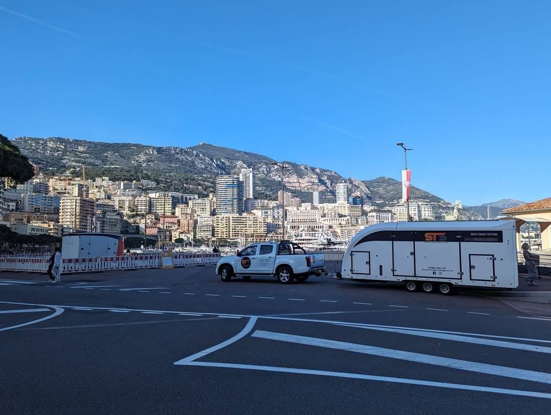 A white truck towing a trailer on a road in Monaco with buildings and hills in the background under a blue sky.