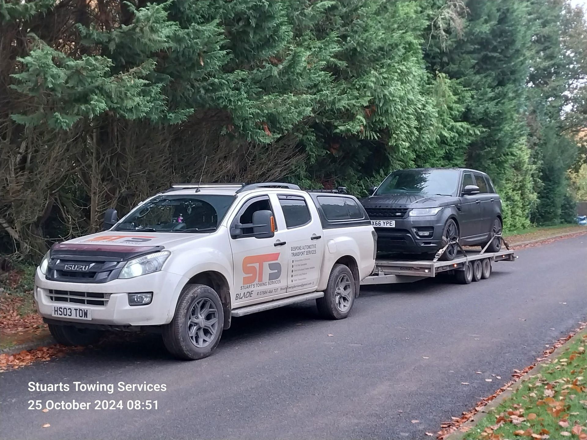 White tow truck towing a black SUV on a trailer along a road, trees in the background.