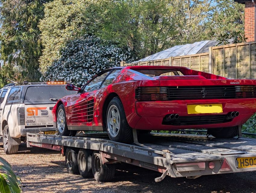 Red Ferrari Testarossa on a trailer, being towed by a white truck. Sunny outdoor setting.
