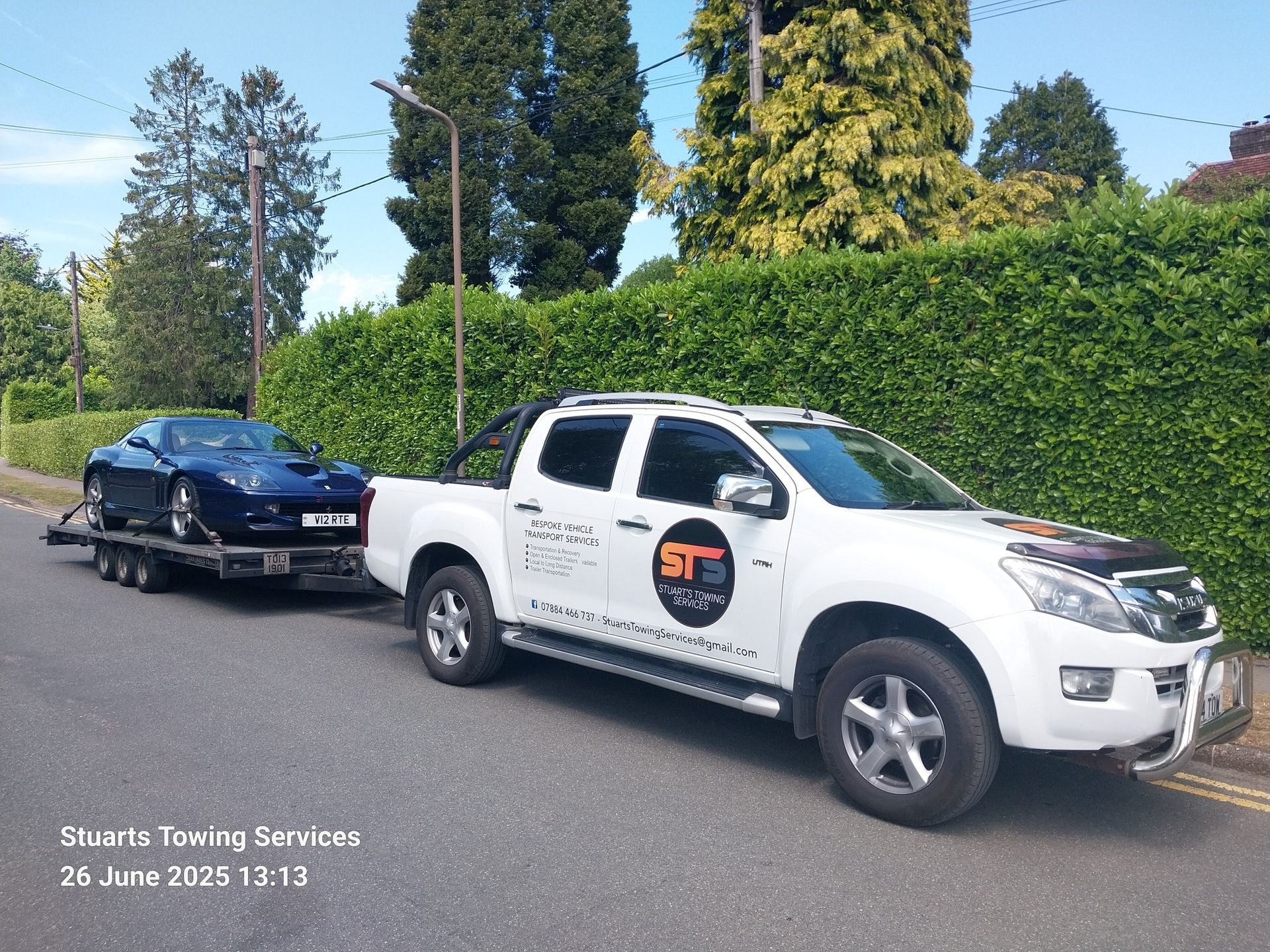 A white truck towing a blue sports car on a trailer; parked on a road, date stamp visible.