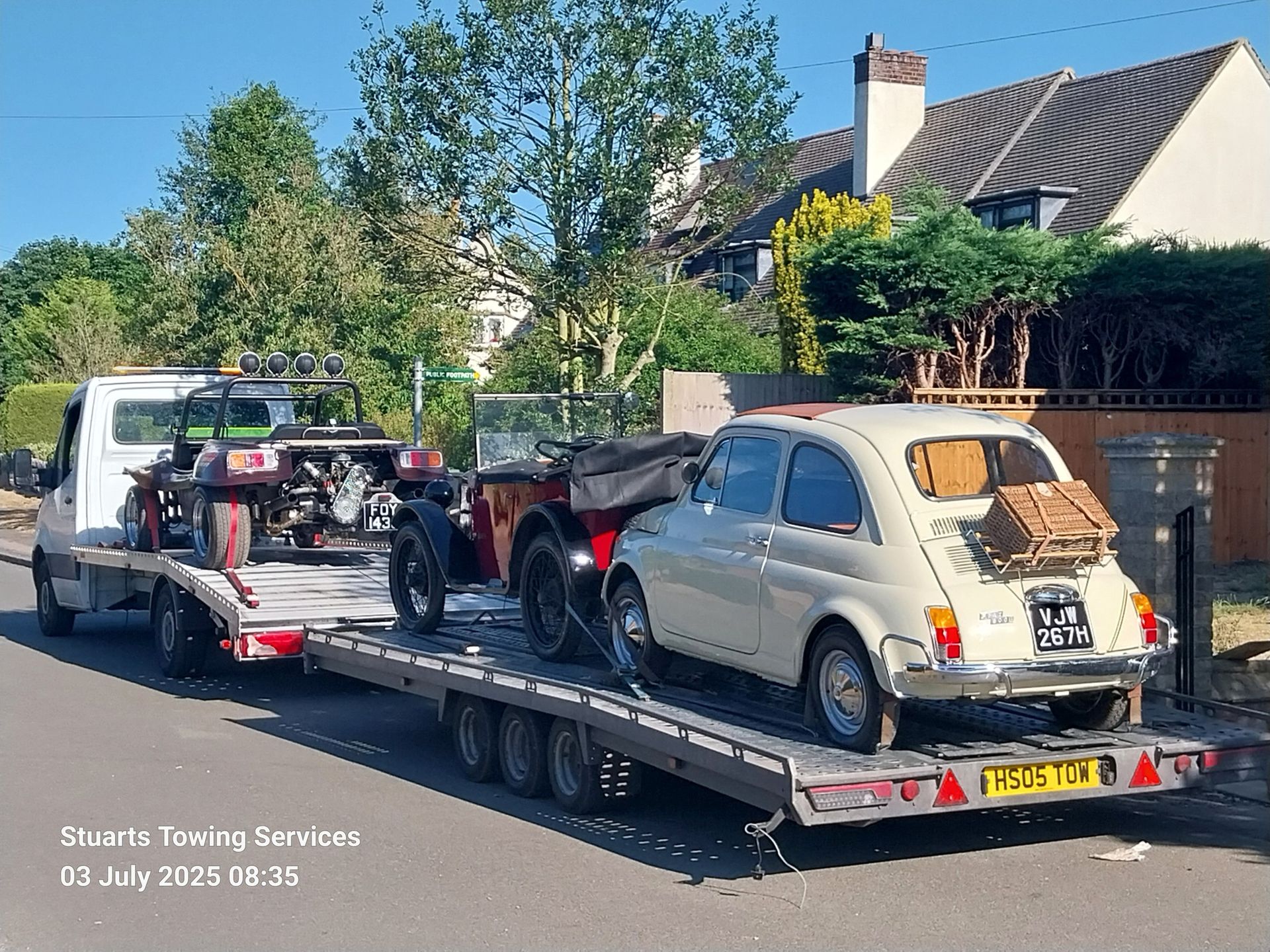 A tow truck carrying two classic cars: a cream Fiat 500 and a vintage sports car.