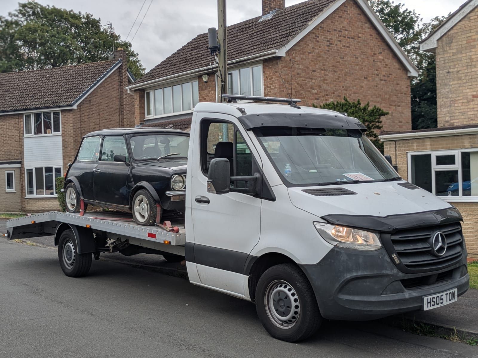 Black Mini Cooper being transported on a flatbed tow truck in front of residential houses.