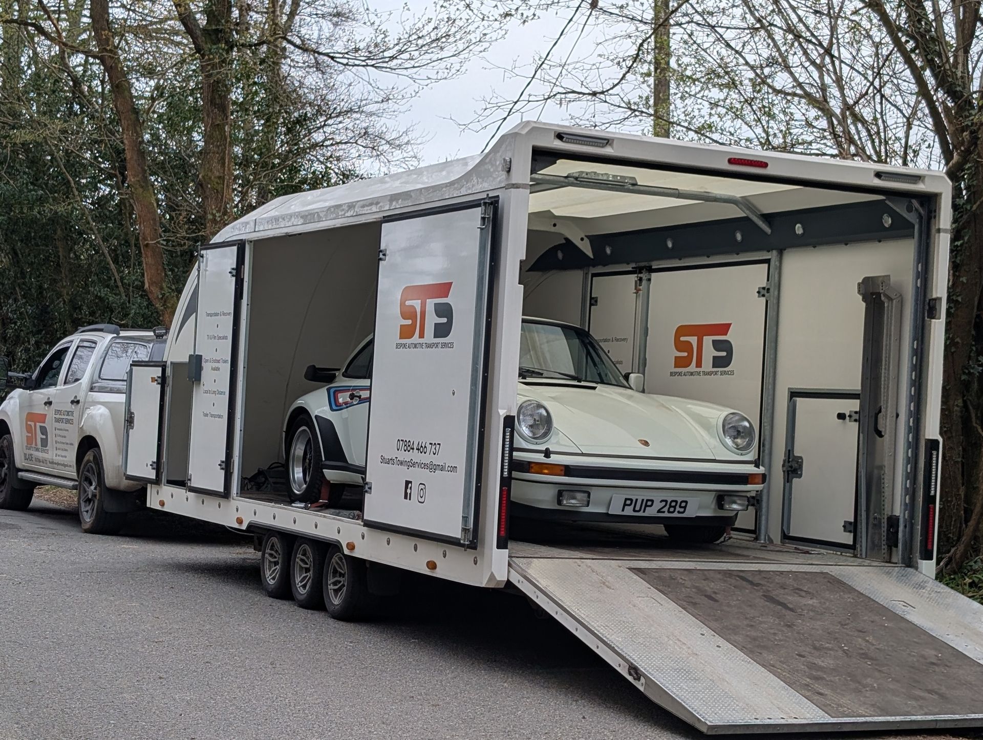 White Porsche inside a car transport trailer being towed by a white pickup truck on a road.