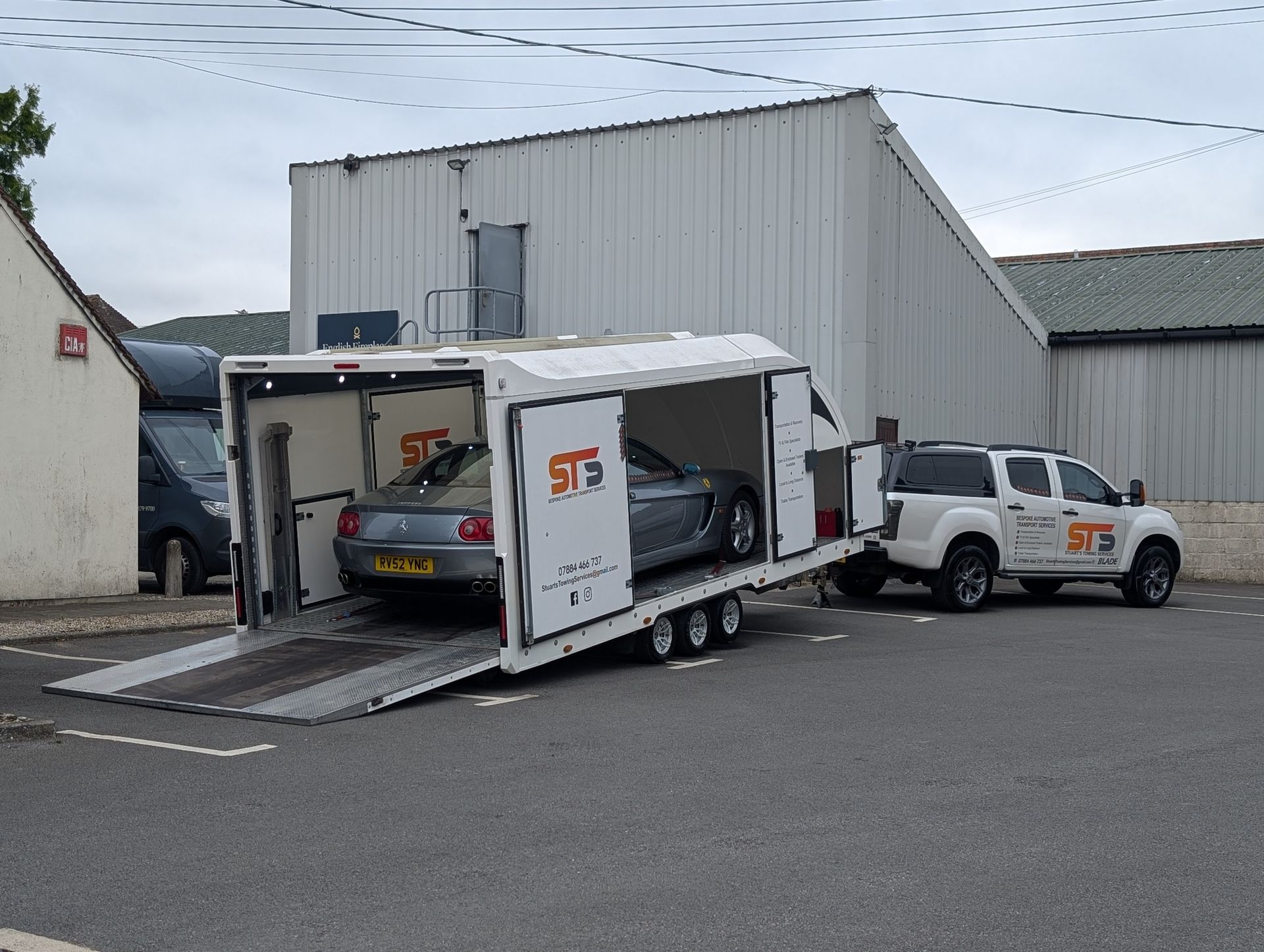 White trailer with car being loaded by truck, parked in front of white building.