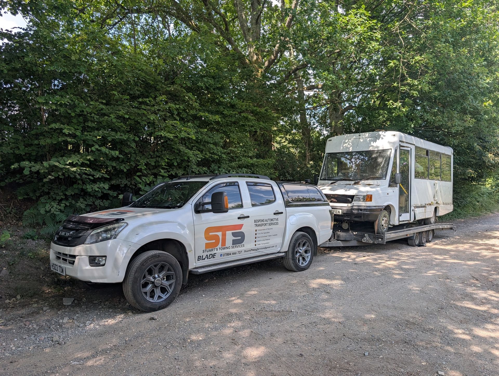 White pickup truck towing a white shuttle bus on a gravel road near a wooded area.