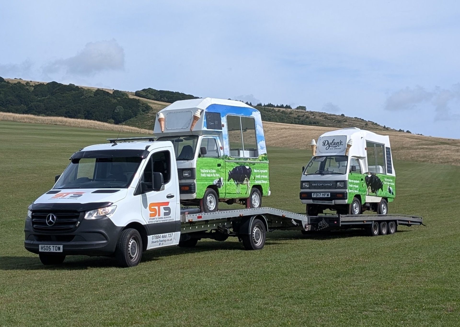 White truck hauling two small buses on a trailer in a grassy field.