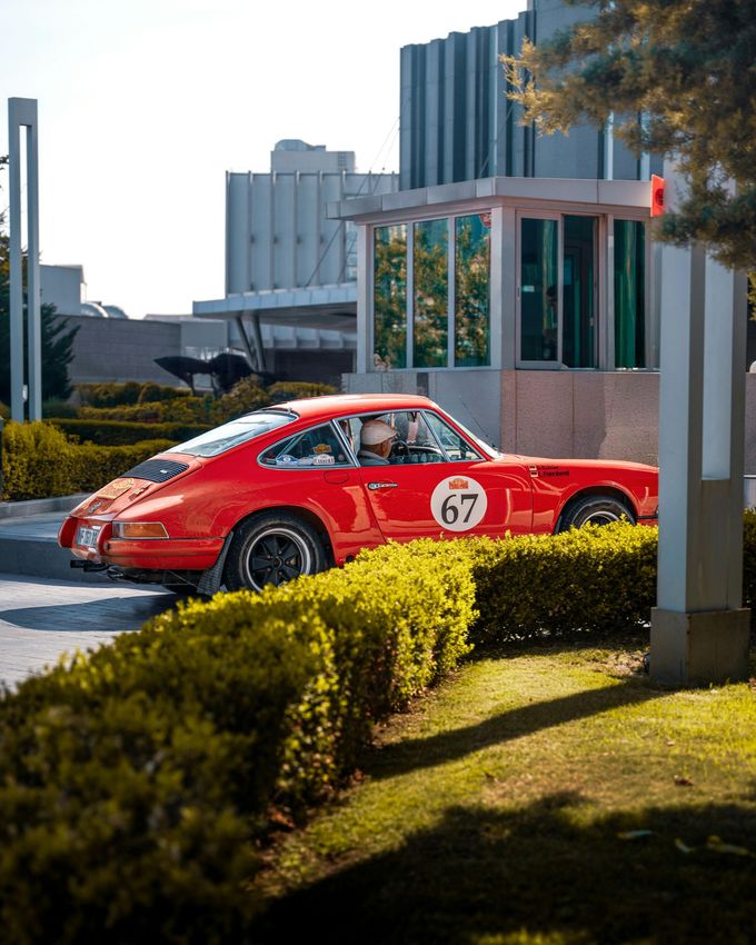 Red classic Porsche 911 with race number 67 driving near a building entrance, sunny day.