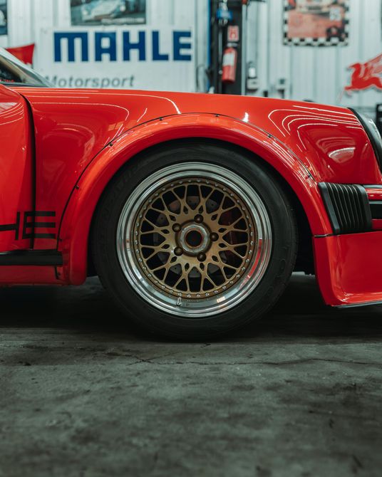 Red Porsche car with gold-rimmed wheel, in a garage setting.