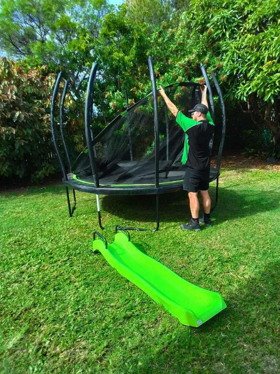 Man Assembling a Trampoline With a Green Slide in a Grassy Yard — Little Green Truck Bundaberg In Maryborough, QLD