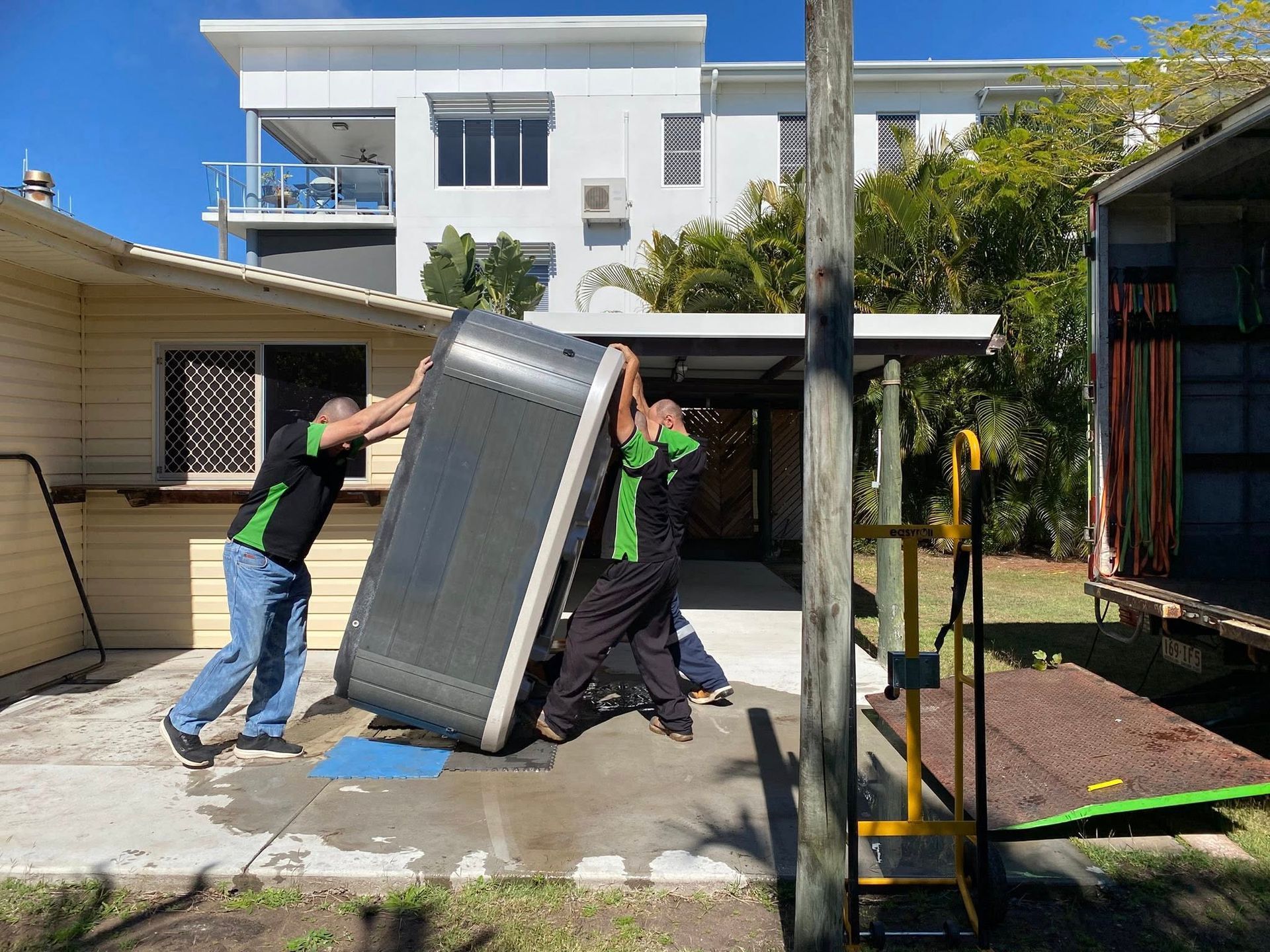 Movers in Green and Black Shirts Lift a Large Dark Object — Little Green Truck Bundaberg In Bundaberg, QLD