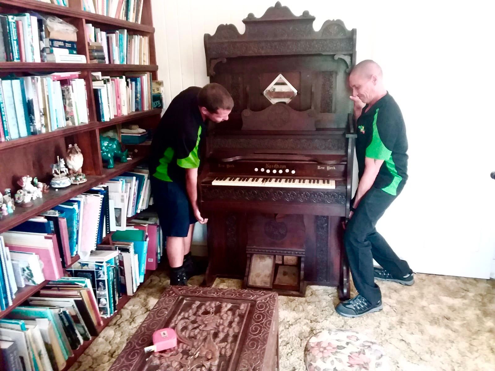 Two people lifting a dark wooden organ in a room with a bookshelf and carpet. - Little Green Truck Bundaberg In Hervey Bay, QLD