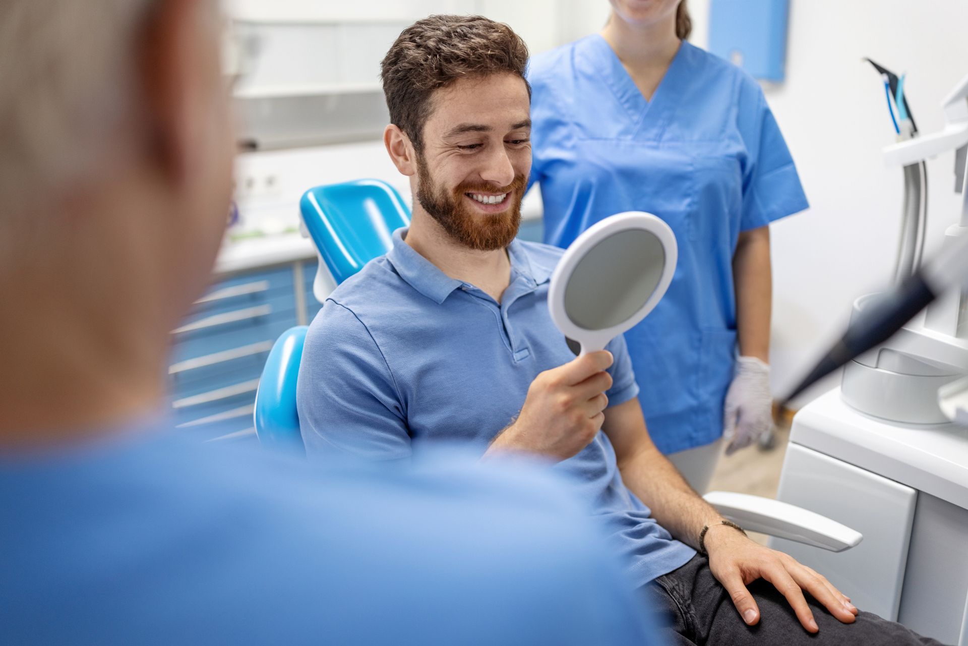 Patient holding a mirror in a dental clinic with two dental professionals nearby. Patient holding a mirror in a dental clinic with two dental professionals nearby.