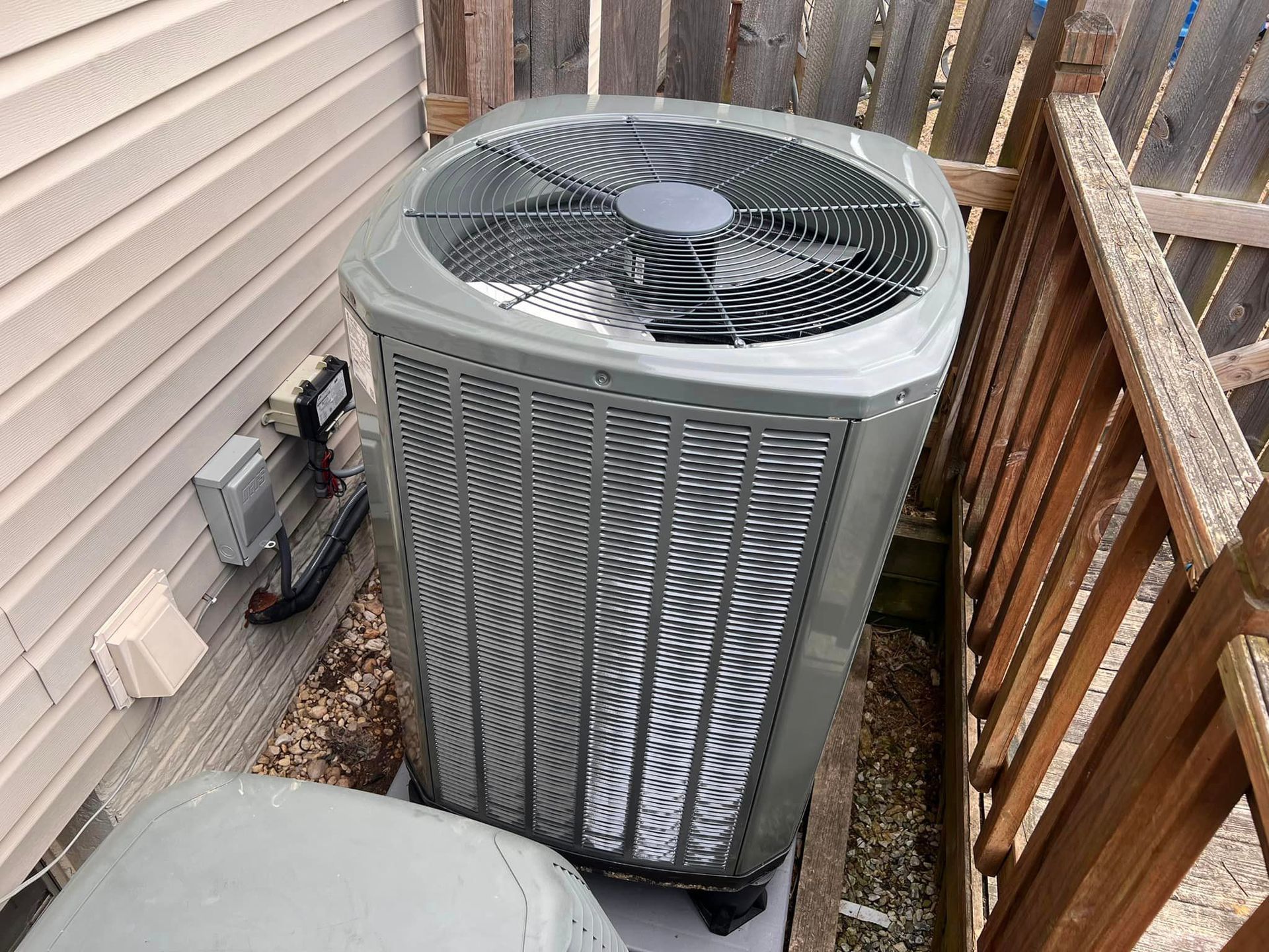 A large air conditioner is sitting on the side of a house next to a wooden fence.