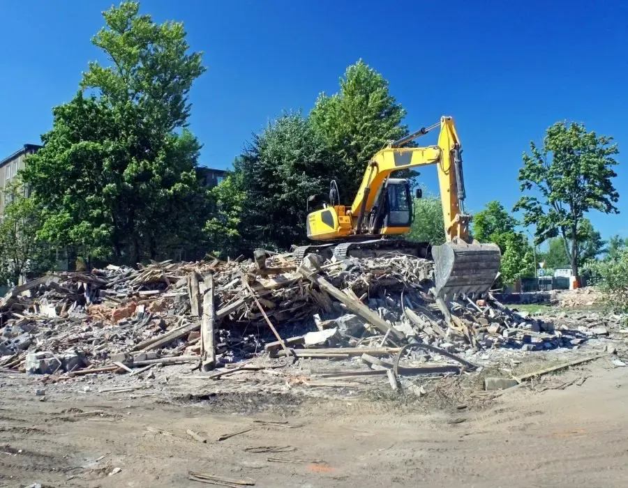 Yellow excavator clearing a pile of rubble at a demolition site with trees and buildings nearby
