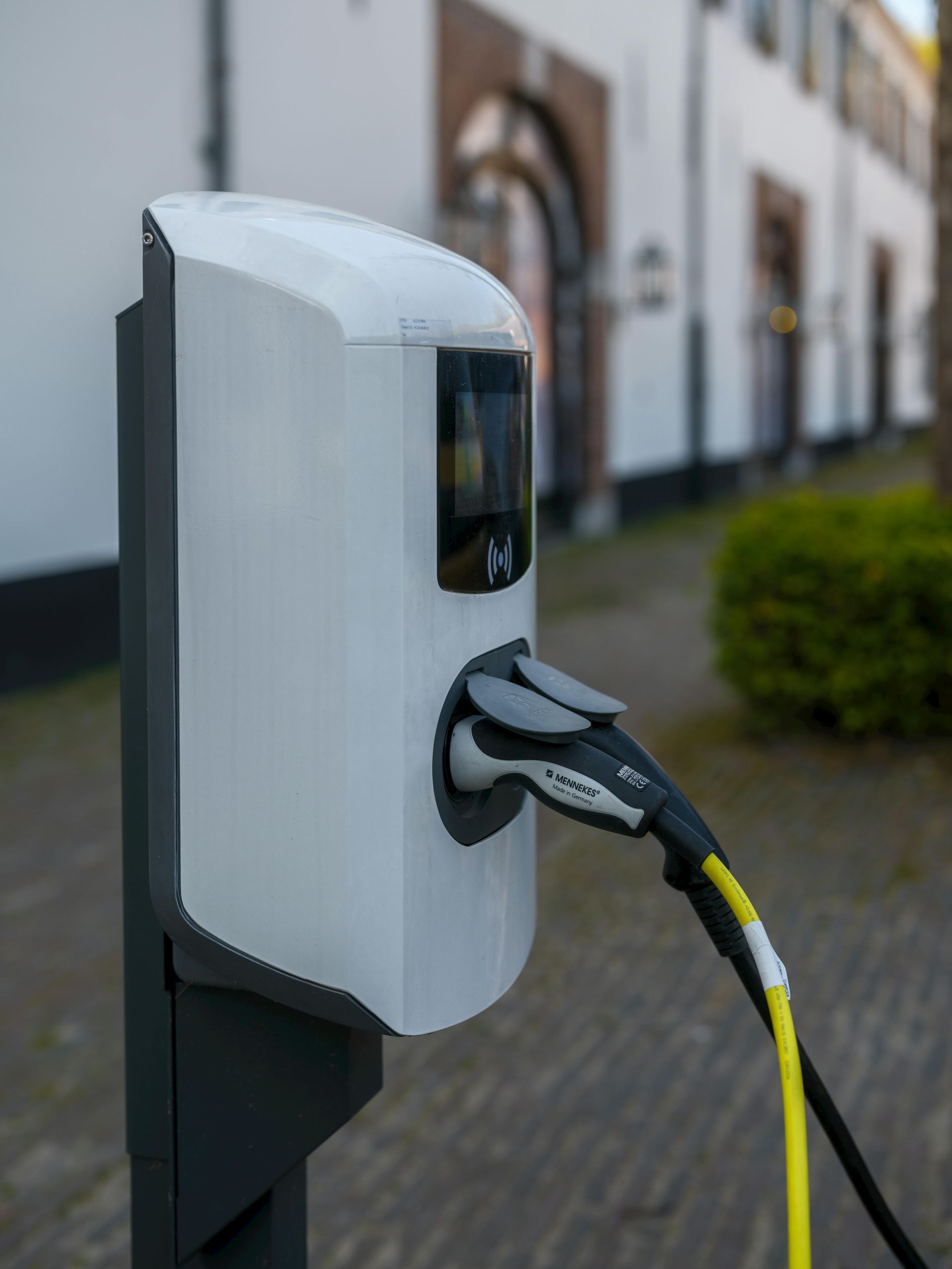 White electric vehicle charging station with plugged-in cable, outdoors near a white building.