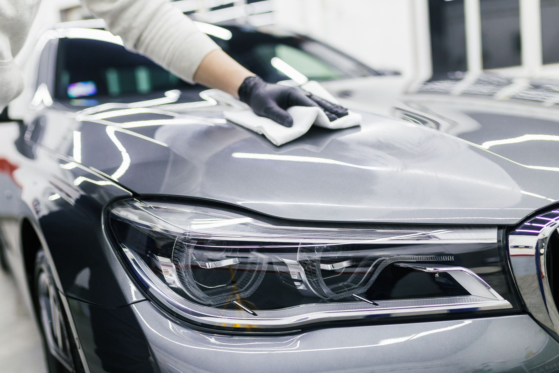 A person in gloves wiping a gray car's hood with a white cloth.
