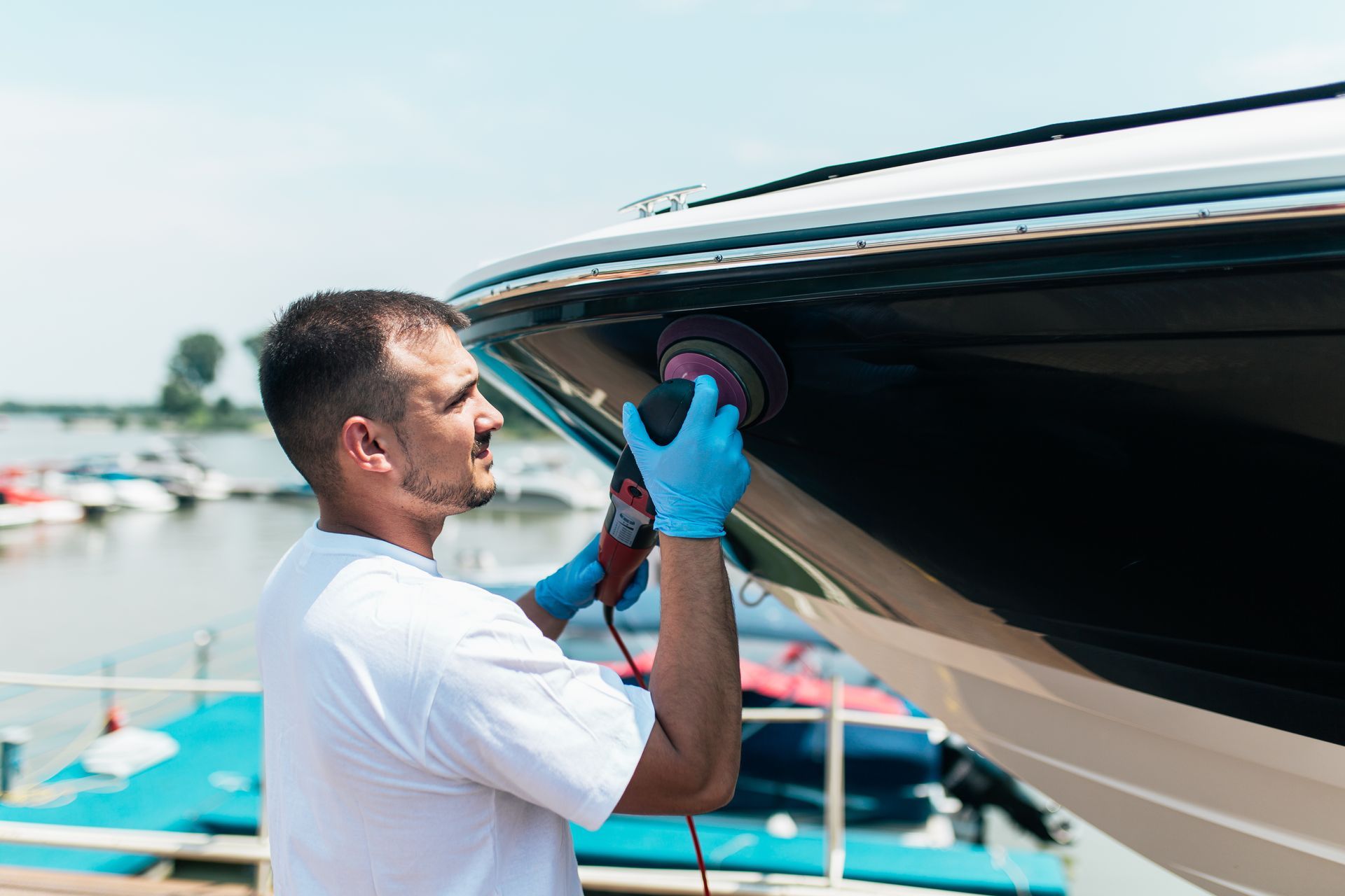Man polishing a boat's hull with a power tool near a dock. He wears gloves and a white shirt.