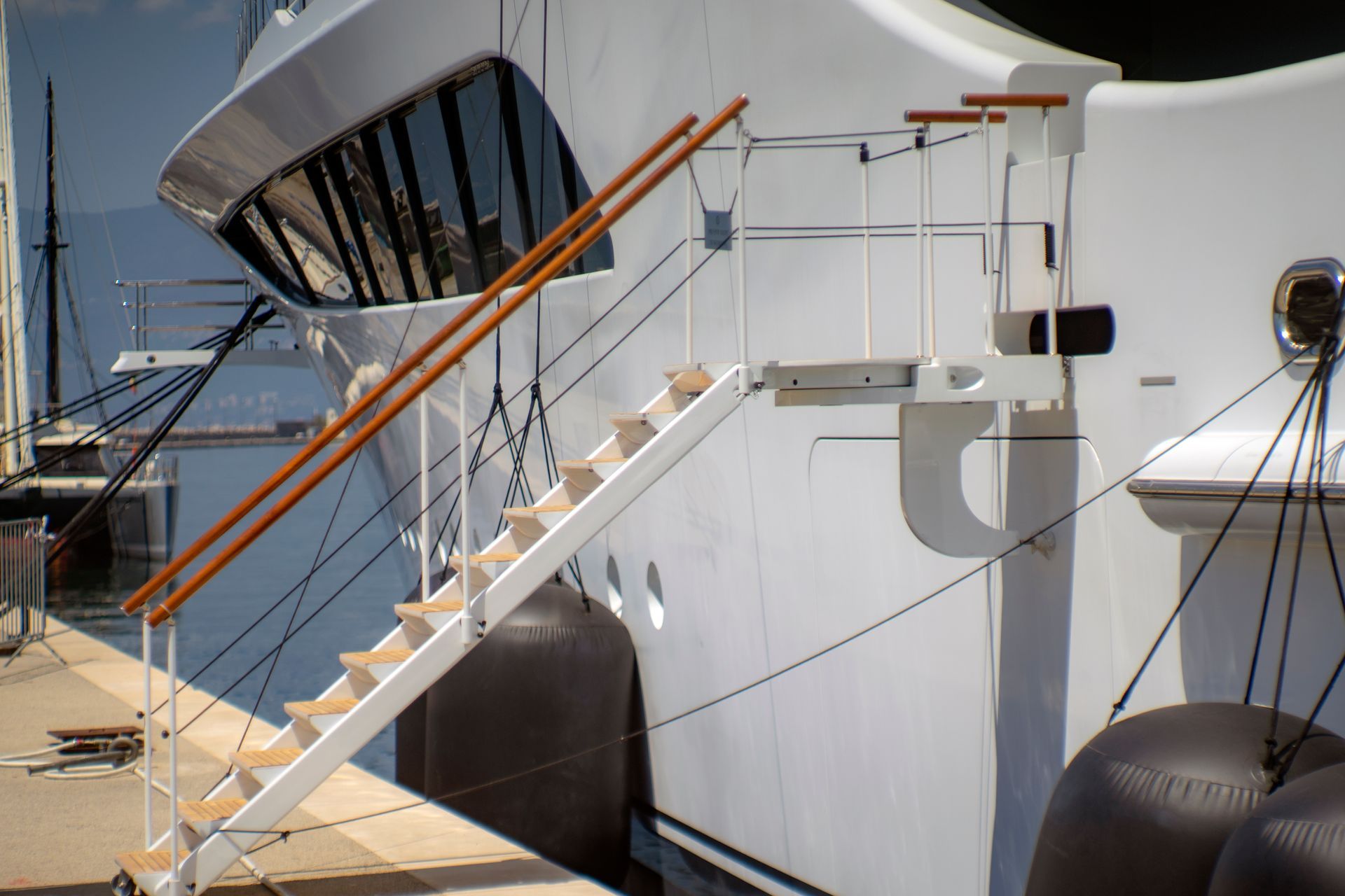 Wooden stairs leading up to a white yacht. Two brown handrails with white supports and a fender are visible.