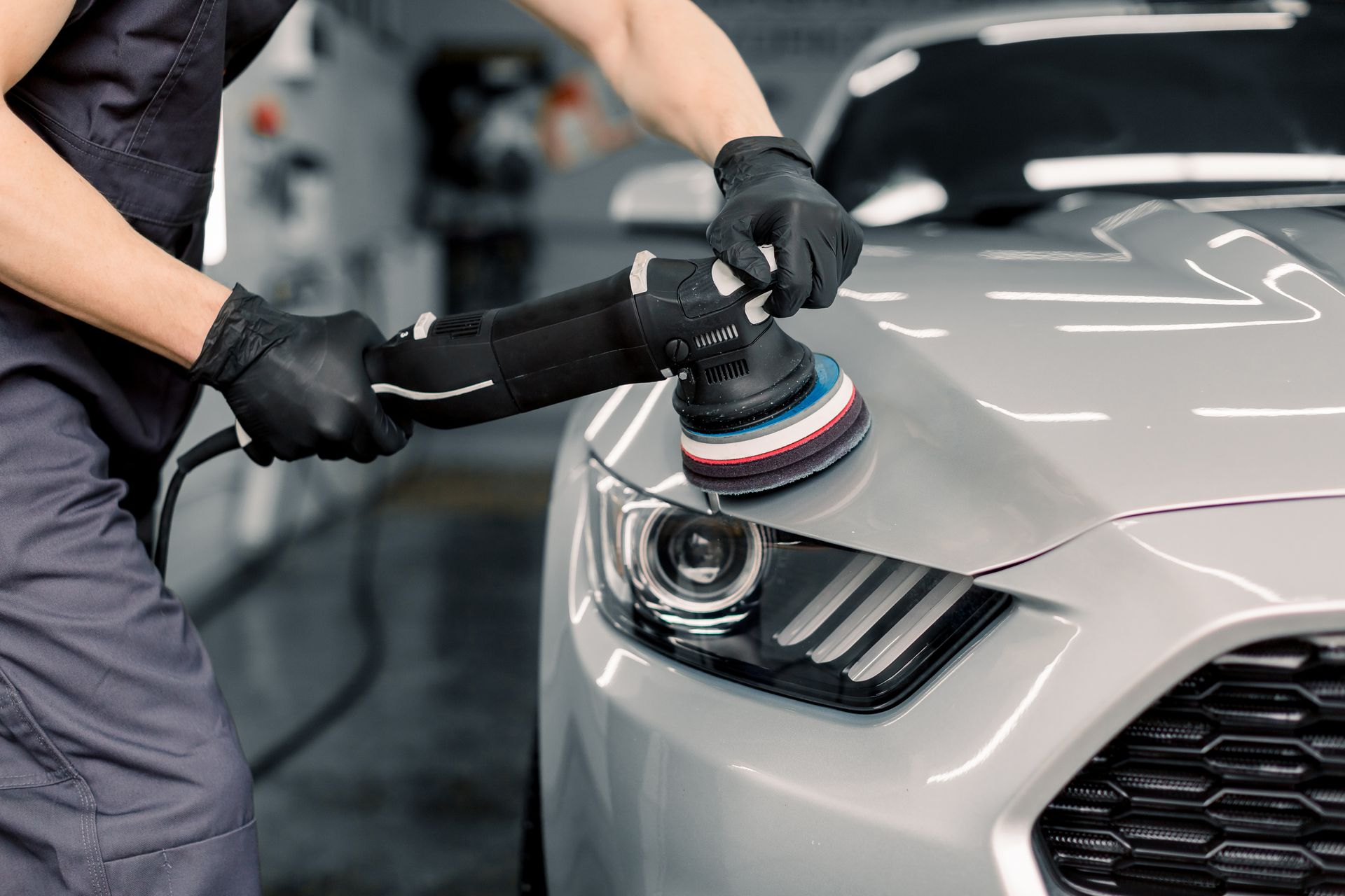 Person using a polisher on a silver car's hood, wearing black gloves. Automotive repair setting.