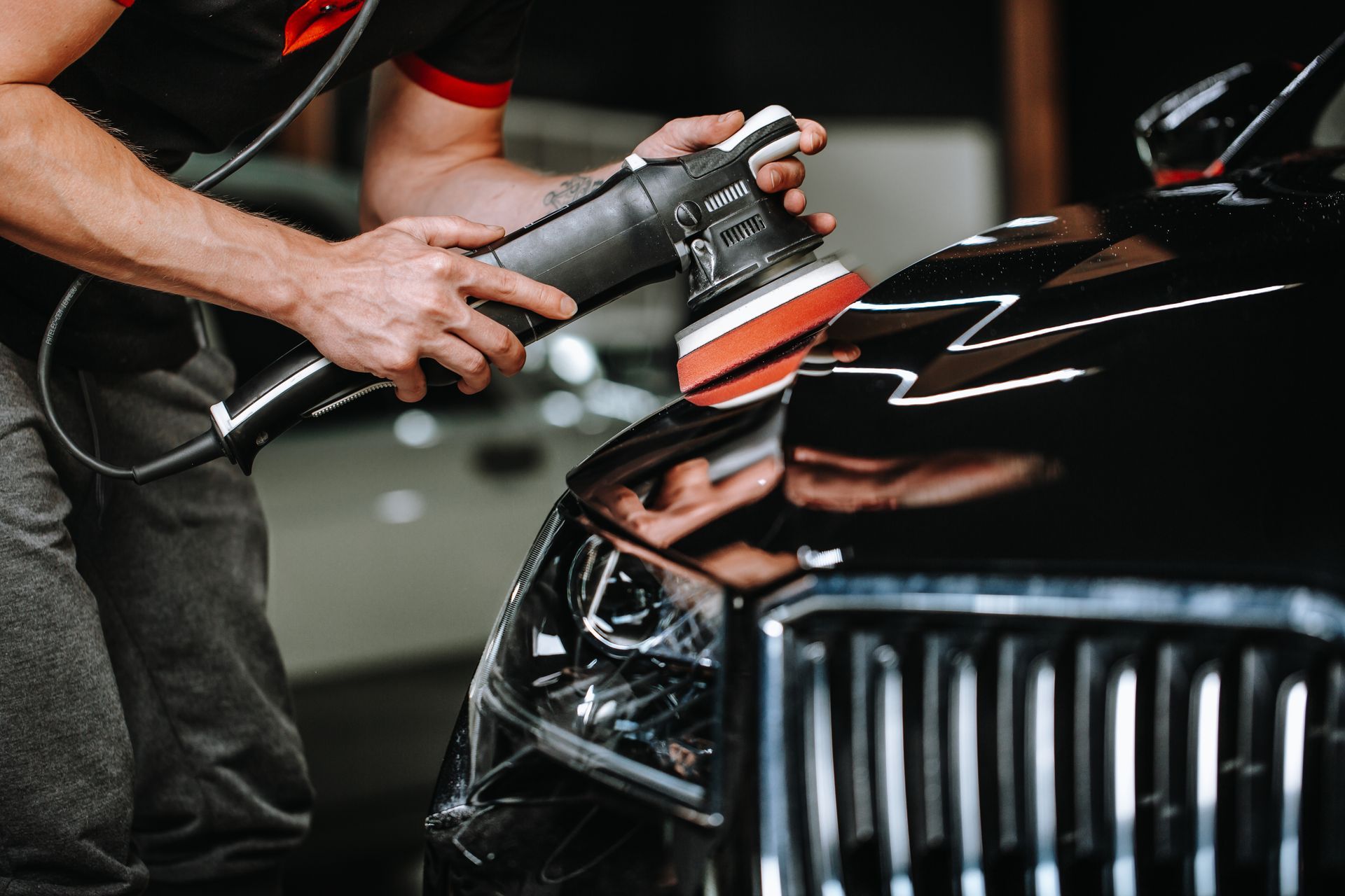 Man polishing a black car hood with an electric buffer in a garage.