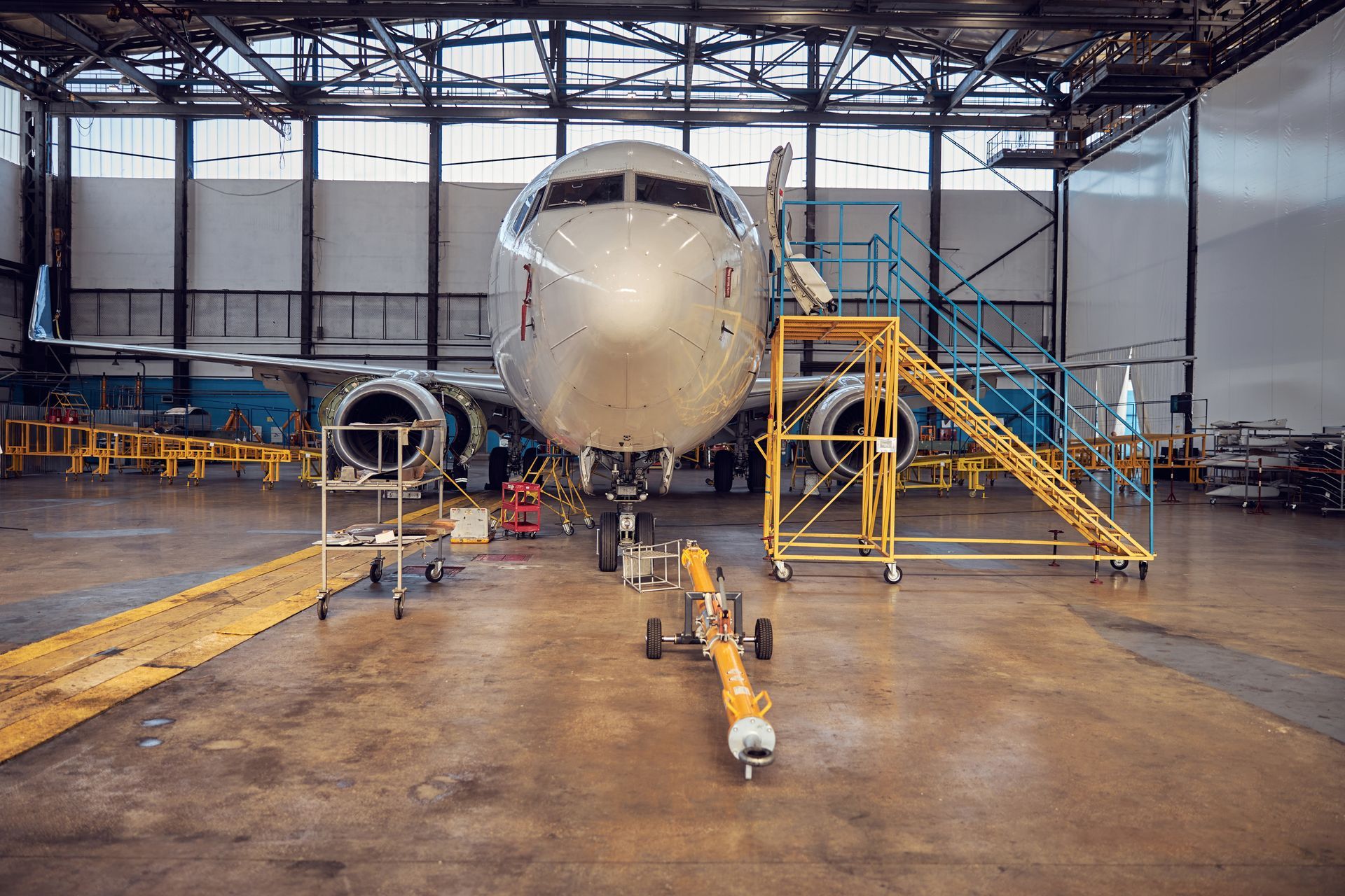 Airplane inside a maintenance hangar, with yellow access stairs and tow bar, on a concrete floor.